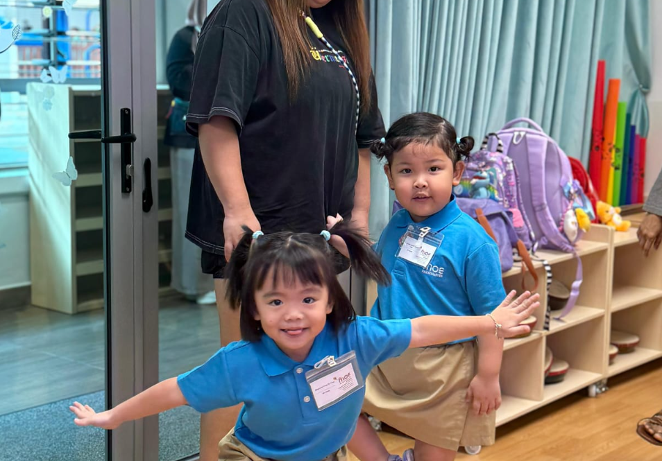 Two young children in blue uniforms pose with arms out as an adult stands behind in a classroom.